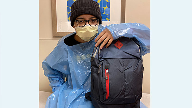 A young boy sits with a blue smock holding a black and red backpack. 